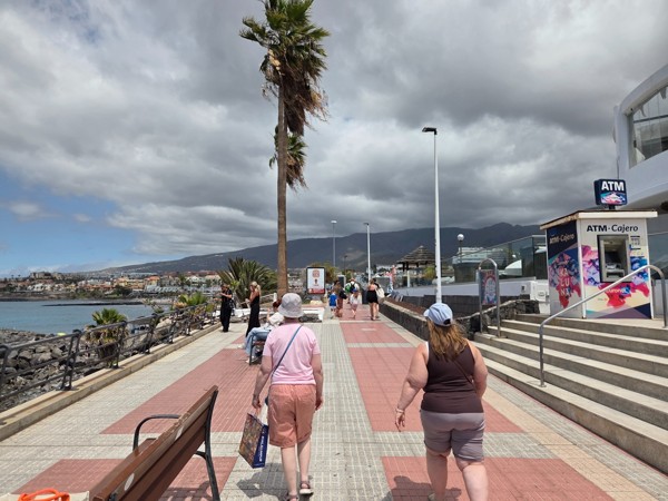 Paseo marítimo de Costa Adeje con personas caminando, palmeras, cajero automático y vista del mar y montañas.