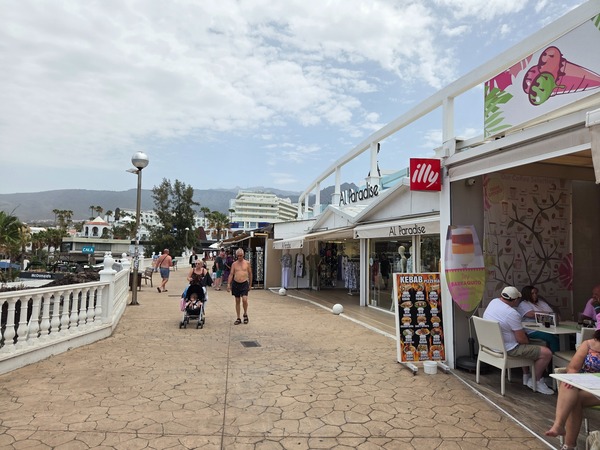 Zona comercial y paseo en Costa Adeje con terrazas, tiendas y vistas hacia los edificios y montañas.
