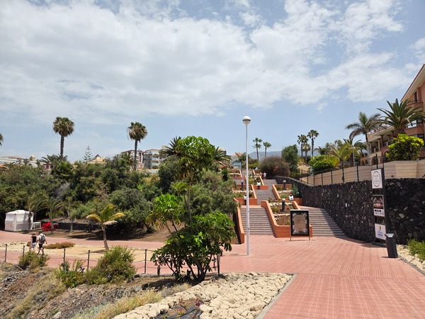 Escaleras y paseo peatonal en Costa Adeje rodeados de palmeras, jardines y edificios residenciales bajo un cielo parcialmente nublado.