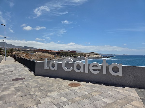 Letrero de La Caleta en un paseo junto al océano con acantilados, costa rocosa y cielo despejado.