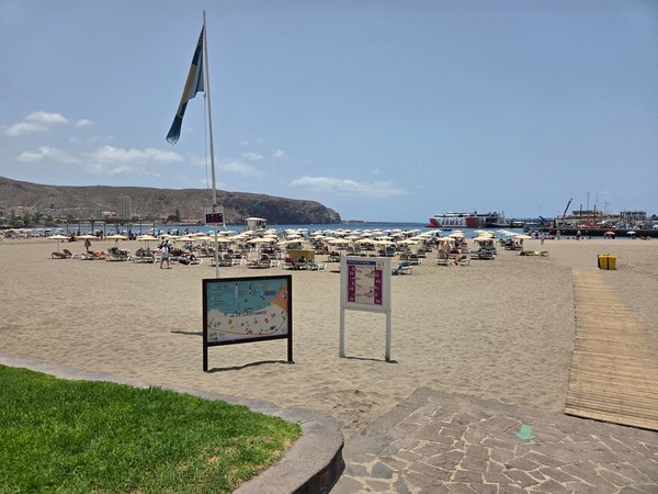 Playa de Los Cristianos con arena clara, tumbonas, sombrillas y el puerto visible al fondo.