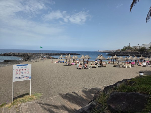 Playa El Bobo con sombrillas de paja, hamacas, bandera verde y una pequeña cala protegida por rocas.